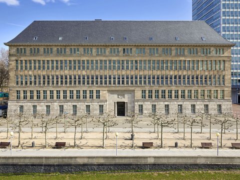 Fassade eines mehrstöckigen historischen Gebäudes mit symmetrischen Fenstern und einem zentralen Eingang, davor ein gepflasterter Platz mit kahlen Bäumen