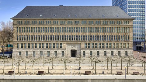 Fassade eines mehrstöckigen historischen Gebäudes mit symmetrischen Fenstern und einem zentralen Eingang, davor ein gepflasterter Platz mit kahlen Bäumen