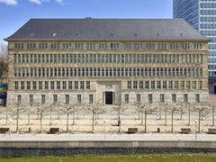 Fassade eines mehrstöckigen historischen Gebäudes mit symmetrischen Fenstern und einem zentralen Eingang, davor ein gepflasterter Platz mit kahlen Bäumen