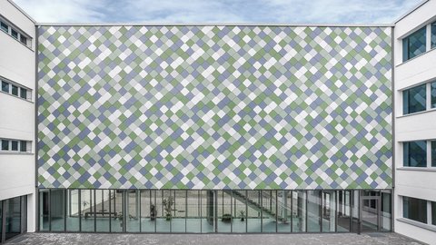 Fassade eines modernen Schulgebäudes mit großflächiger, quadratischer Kachelverkleidung in einem diagonalen Muster und bodentiefen Fenstern im Erdgeschoss