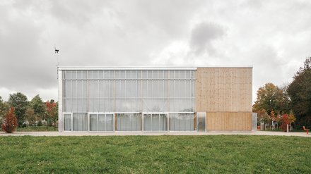 Lange Gebäudeansicht: zweigeschossige Glasfront mit Vorhängen links, geschlossene Holzverkleidung rechts; Wiese davor.