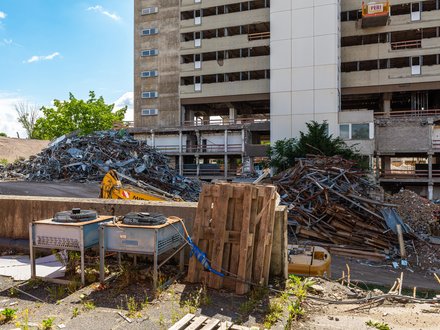 Foto einer Rückbau‑Baustelle: teilentkerntes Hochhaus, vorne Holzpaletten und Geräte; links und rechts große Haufen aus Schrottmetall und Ziegeln, Baggerarm sichtbar; sonniger Tag, vereinzelt Bewuchs am Rand.