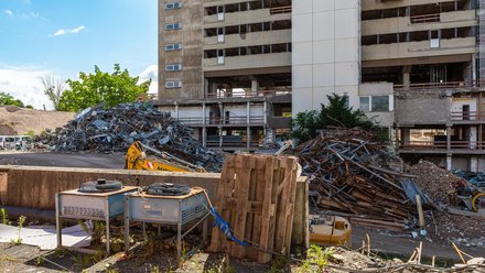 Foto einer Rückbau‑Baustelle: teilentkerntes Hochhaus, vorne Holzpaletten und Geräte; links und rechts große Haufen aus Schrottmetall und Ziegeln, Baggerarm sichtbar; sonniger Tag, vereinzelt Bewuchs am Rand.