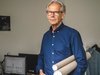 Mann in blauem Hemd hält zwei Papprollen und steht in einem Büro mit Computer und Kleidung im Hintergrund.