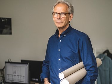 Mann in blauem Hemd hält zwei Papprollen und steht in einem Büro mit Computer und Kleidung im Hintergrund.