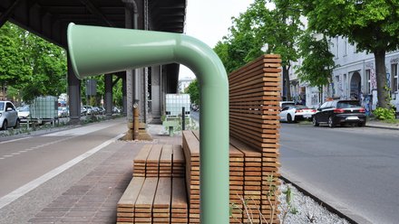 Sitzbank und Stadtmöbel entlang des Radwegs unter der Hochbahn, mit Holzmodulen, Begrünung und Straße im Hintergrund.