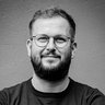 "Porträt eines Mannes mit Vollbart und randloser Brille, schwarzem T-Shirt, die Arme über der Brust verschränkt. Der Mann lächelt in die Kamera vor grauem Hintergrund."