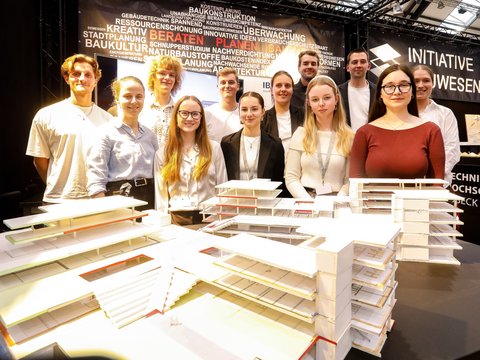 Eine Gruppe junger Erwachsener steht dicht hinter mehreren detaillierten Architekturmodellen auf einem Ausstellungstisch und blickt gemeinsam in die Kamera, der Messestand ist von Fachbegriffen zu Architektur, Planung und Bauwesen umgeben, wodurch das Bild Teamarbeit, Ausbildung und gemeinsames Arbeiten an architektonischen Konzepten sichtbar macht und für kollektives Lernen und Nachwuchs im Bauwesen steht.