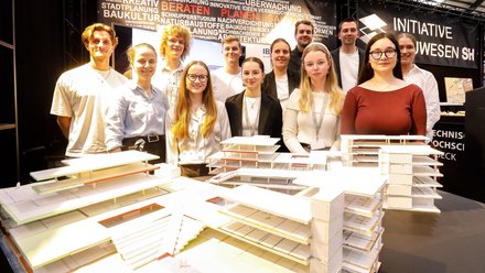 Eine Gruppe junger Erwachsener steht dicht hinter mehreren detaillierten Architekturmodellen auf einem Ausstellungstisch und blickt gemeinsam in die Kamera, der Messestand ist von Fachbegriffen zu Architektur, Planung und Bauwesen umgeben, wodurch das Bild Teamarbeit, Ausbildung und gemeinsames Arbeiten an architektonischen Konzepten sichtbar macht und für kollektives Lernen und Nachwuchs im Bauwesen steht.