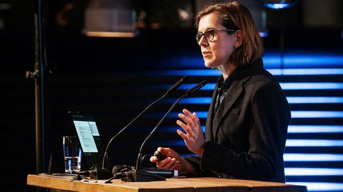 Das Bild zeigt eine Person an einem Rednerpult, die offenbar gerade einen Vortrag hält. Vor der Person stehen zwei Mikrofone, auf dem Pult liegt ein Laptop. Die Person gestikuliert mit einer Hand, was auf eine erklärende oder argumentierende Rede hinweist. Im Hintergrund sind Lichtinstallationen und Teile des Veranstaltungsraums zu sehen. Zweck des Bildes ist es, einen Vortrag oder eine Keynote im Rahmen der Veranstaltung zu dokumentieren und den Moment der Wissensvermittlung oder Ansprache festzuhalten.