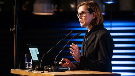 Das Bild zeigt eine Person an einem Rednerpult, die offenbar gerade einen Vortrag hält. Vor der Person stehen zwei Mikrofone, auf dem Pult liegt ein Laptop. Die Person gestikuliert mit einer Hand, was auf eine erklärende oder argumentierende Rede hinweist. Im Hintergrund sind Lichtinstallationen und Teile des Veranstaltungsraums zu sehen. Zweck des Bildes ist es, einen Vortrag oder eine Keynote im Rahmen der Veranstaltung zu dokumentieren und den Moment der Wissensvermittlung oder Ansprache festzuhalten.