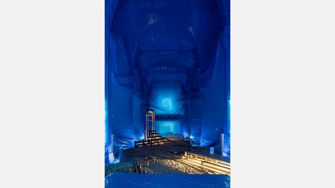 Kirchenraum mit blau verhüllten Wänden und Decken. Ein sandbedeckter Boden mit Holzstegen führt durch die Installation.