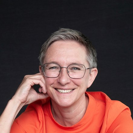 Porträt einer Person mittleren Alters mit grauem kurzem Haar und Brille. Sie trägt ein leuchtend orangerotes T-Shirt und sitzt vor einem dunklen Hintergrund. Mit der linken Hand gestützt auf den Tisch lächelt sie freundlich in die Kamera. Sie trägt eine silberne Uhr und einen Ring an der linken Hand.