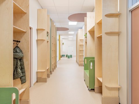 "Langer Flur einer Kindereinrichtung mit beidseitig angeordneten Holzfächern aus hellem Sperrholz. Links hängt eine schwarze Jacke an einem Haken. Jedes Fachmodul hat mehrere offene Ablageflächen. Grüne Kunststoff-Behälter stehen im hinteren Bereich. Weiße Decke mit indirekter Beleuchtung und ovalen Akzenten in Braun und Creme. Heller Bodenbelag."