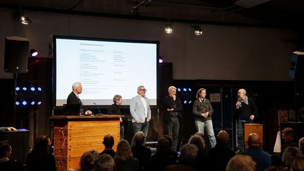 Das Bild zeigt eine Bühne in einem Veranstaltungsraum. Mehrere Personen stehen nebeneinander vor einem großen Bildschirm, auf dem eine Präsentation mit Text angezeigt wird. Im Vordergrund ist das Publikum zu sehen, das auf die Bühne blickt. Die Situation wirkt wie eine Podiumsdiskussion, Vorstellung oder Preisverleihung. Zweck des Bildes ist es, den formellen Teil einer Veranstaltung zu zeigen und den Austausch oder die Präsentation von Inhalten vor Publikum zu dokumentieren.