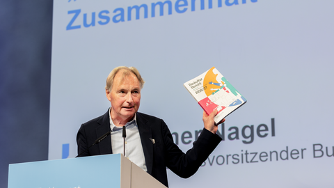 „Ein Mann spricht an einem Rednerpult beim Konvent der Baukultur und hält den Bericht „Baukultur Bericht 2026/27“ in die Höhe. Auf der Projektionsfläche im Hintergrund steht unter anderem „Gestalt Zusammenhalt“.“