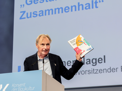 „Ein Mann spricht an einem Rednerpult beim Konvent der Baukultur und hält den Bericht „Baukultur Bericht 2026/27“ in die Höhe. Auf der Projektionsfläche im Hintergrund steht unter anderem „Gestalt Zusammenhalt“.“