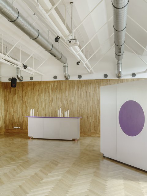 Ein großer Freizeitraum mit hellem Holzparkettboden. Die Rückwand ist mit vertikalen Holzlatten verkleidet. Im Hintergrund steht ein grauer Tresen auf Rollen, auf dem leere Becher stehen. Links ist eine Dartscheibe und ein Lautsprecher an der Wand befestigt. Rechts im Vordergrund steht eine weiße Trennwand mit einem großen lila Kreis. An der Decke sind silberne Lüftungsrohre und Strahler sichtbar.