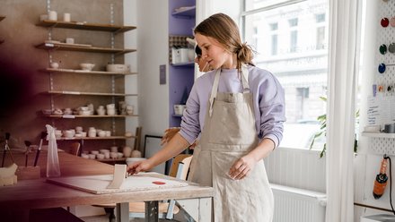 13:02     Person mit Schürze formt an einer Tonplatte auf einer Werkbank in heller Keramikwerkstatt; im Hintergrund Regale mit Schalen, seitlich fällt Tageslicht durchs Fenster.