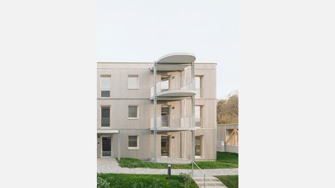 Wohngebäude mit heller Holzfassade und übereinanderliegenden Balkonen, davor Rasen und gepflasterter Zugang.