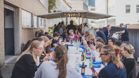 Menschen sitzen an langen Tischen im Freien und essen oder unterhalten sich in einer urbanen Umgebung