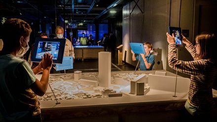 "Ausstellungsbereich mit einem weißen Stadtmodell aus einfachen geometrischen Formen. Kinder und Erwachsene nutzen Tablets, um digitale Inhalte über das Modell zu erkunden. Zwei Kinder halten die Geräte hoch und richten sie auf das Modell. Weitere Besucher stehen im Hintergrund. Der Raum ist gedämpft beleuchtet, mit Fokus auf die interaktive Station
