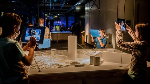"Ausstellungsbereich mit einem weißen Stadtmodell aus einfachen geometrischen Formen. Kinder und Erwachsene nutzen Tablets, um digitale Inhalte über das Modell zu erkunden. Zwei Kinder halten die Geräte hoch und richten sie auf das Modell. Weitere Besucher stehen im Hintergrund. Der Raum ist gedämpft beleuchtet, mit Fokus auf die interaktive Station