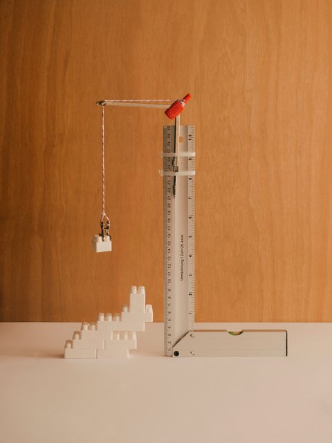 "Minimalistische Illustration eines Baukrans vor orangebraunem Hintergrund. Ein weißer Messstab dient als Kranarm mit rotem Haken, der einen weißen Block hebt. Am Boden sind gestaffelte weiße Bauklötze angeordnet. Symbolisiert Bauwirtschaft oder Produktivität."