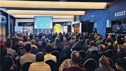 "Vollbesetzter Veranstaltungsraum mit blauem Wand- und Deckendesign. Im Vordergrund sitzen etwa hundert Besucherinnen und Besucher in Stuhlreihen, überwiegend mittleren und älteren Alters, in verschiedenfarbiger Kleidung. Vorne auf der Bühne präsentiert ein Referent unter einer Leinwand mit türkisem Hintergrund. Gelbe und orangefarbene Informationstafeln, Fahrradausstellung und weitere Ausstellungselemente sind sichtbar. Warme Deckenbeleuchtung erhellt den Raum."