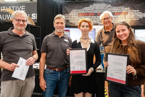 Mehrere Personen zeigen Urkunden und Anerkennungen auf einem Messestand.
