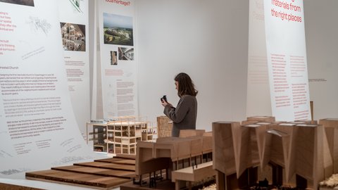 "Heller Ausstellungsraum mit großen weißen Informationstafeln, die Texte und Bilder zu Architekturprojekten zeigen. Eine Besucherin steht vor einem Tisch mit detaillierten Holzmodellen von Gebäuden und fotografiert eines der Modelle mit ihrem Smartphone. Der Raum wirkt ruhig und minimalistisch gestaltet."