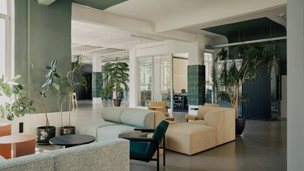 Heller Büro‑Loungebereich mit modularen Sofas in Grün und Beige, niedrigen Tischen und vielen großen Zimmerpflanzen; verglaste Besprechungsräume im Hintergrund.