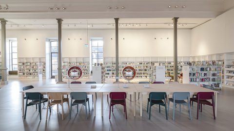 Großer heller Lesesaal mit langem weißen Tisch und bunten Stühlen vor Bücherregalen und großen Fenstern