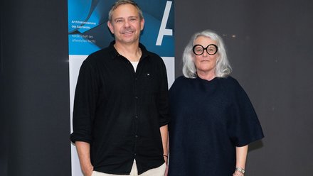 Zwei erwachsene Personen stehen nebeneinander vor demselben Banner, beide lächeln leicht und sind frontal zur Kamera ausgerichtet, was das Bild als formelles Porträt innerhalb eines institutionellen Rahmens kennzeichnet und für Leitung, Repräsentation und Kontinuität innerhalb einer Organisation steht.