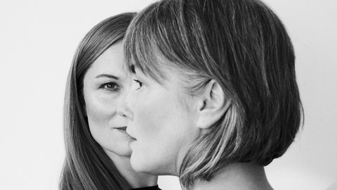 "Schwarzweiß-Porträtfoto zweier Frauen von hinten, überlagert in doppelter Belichtung. Beide haben schulterlange Haare mit Pony. Eine Frau schaut nach links, die andere nach rechts. Beide tragen dunkle Kleidung vor neutralem, hellem Hintergrund."