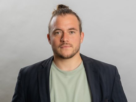 Porträtfoto eines jungen Mannes mit braunem Haar zu einem Dutt gebunden und dunklem Bart. Er trägt einen dunkelblauen Blazer über einem hellgrünen T-Shirt und blickt direkt in die Kamera vor neutralem grauem Hintergrund."