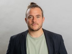 Porträtfoto eines jungen Mannes mit braunem Haar zu einem Dutt gebunden und dunklem Bart. Er trägt einen dunkelblauen Blazer über einem hellgrünen T-Shirt und blickt direkt in die Kamera vor neutralem grauem Hintergrund."