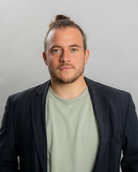 Porträtfoto eines jungen Mannes mit braunem Haar zu einem Dutt gebunden und dunklem Bart. Er trägt einen dunkelblauen Blazer über einem hellgrünen T-Shirt und blickt direkt in die Kamera vor neutralem grauem Hintergrund."