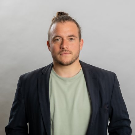 Porträtfoto eines jungen Mannes mit braunem Haar zu einem Dutt gebunden und dunklem Bart. Er trägt einen dunkelblauen Blazer über einem hellgrünen T-Shirt und blickt direkt in die Kamera vor neutralem grauem Hintergrund."