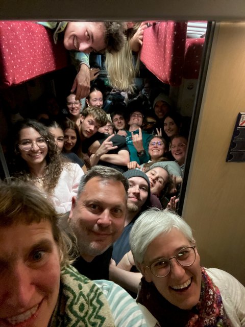 „Eine große, fröhliche Reisegruppe posiert für ein dicht gedrängtes Gruppen-Selfie in einem Zugabteil.“