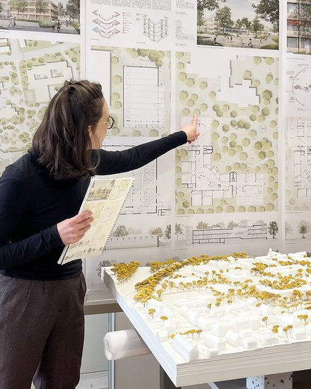 Person präsentiert städtebauliche Entwürfe; zeigt auf große Wandpläne mit Lageplänen und Grundrissen, hält ein Heft; im Vordergrund ein weißes Architekturmodell mit gelben Bäumen auf einem Tisch.