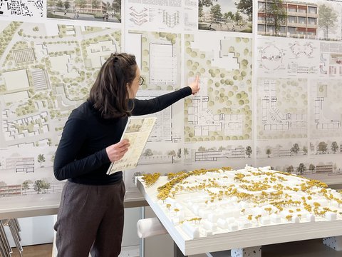 Person präsentiert städtebauliche Entwürfe; zeigt auf große Wandpläne mit Lageplänen und Grundrissen, hält ein Heft; im Vordergrund ein weißes Architekturmodell mit gelben Bäumen auf einem Tisch.