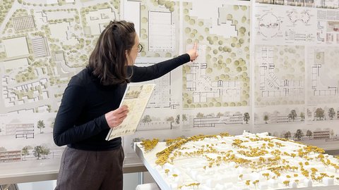 Person präsentiert städtebauliche Entwürfe; zeigt auf große Wandpläne mit Lageplänen und Grundrissen, hält ein Heft; im Vordergrund ein weißes Architekturmodell mit gelben Bäumen auf einem Tisch.