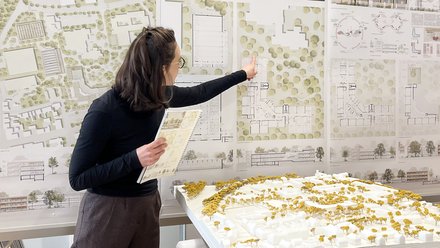 Person präsentiert städtebauliche Entwürfe; zeigt auf große Wandpläne mit Lageplänen und Grundrissen, hält ein Heft; im Vordergrund ein weißes Architekturmodell mit gelben Bäumen auf einem Tisch.