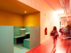 Schulflur mit rotem Boden und Sitznische mit Tisch und Sitzbänken in grüner und gelber Wandfarbe, mehrere Kinder mit Rucksäcken laufen vorbei
