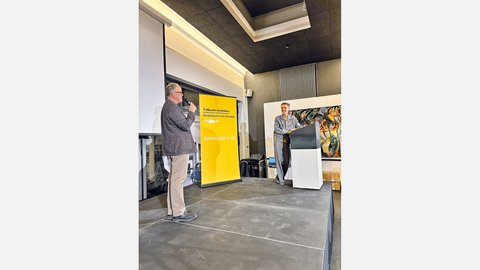 "Konferenzraum mit moderner Architektur: schwarze Decke, helle LED-Leuchten und großes Oberlicht. Links an einem Mikrofon ein Mann mittleren Alters in grauem Pullover und Khaki-Hose. Rechts auf der Bühne ein älterer Mann in hellblauem Hemd, sprechend. Dazwischen eine große gelbe Tafel mit Schriftzug 'Treffpunkt Architektur' und Website. An der rechten Wand ein abstraktes farbiges Kunstwerk. Beton-Fußboden im modernen Stil."