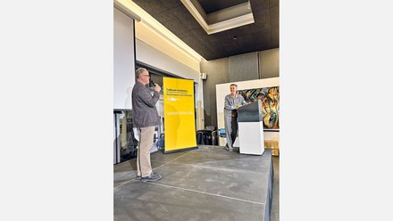 "Konferenzraum mit moderner Architektur: schwarze Decke, helle LED-Leuchten und großes Oberlicht. Links an einem Mikrofon ein Mann mittleren Alters in grauem Pullover und Khaki-Hose. Rechts auf der Bühne ein älterer Mann in hellblauem Hemd, sprechend. Dazwischen eine große gelbe Tafel mit Schriftzug 'Treffpunkt Architektur' und Website. An der rechten Wand ein abstraktes farbiges Kunstwerk. Beton-Fußboden im modernen Stil."