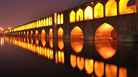 Eine nächtliche Aufnahme zeigt eine lange steinerne Brücke mit regelmäßig gereihten Bögen, die warm beleuchtet sind und sich im ruhigen Wasser spiegeln, die rhythmische Abfolge von Licht und Struktur prägt den Raum, wodurch das Bild historische Ingenieurbaukunst als atmosphärischen Stadtraum zeigt und für Dauerhaftigkeit, Orientierung und symbolische Verbindung steht.