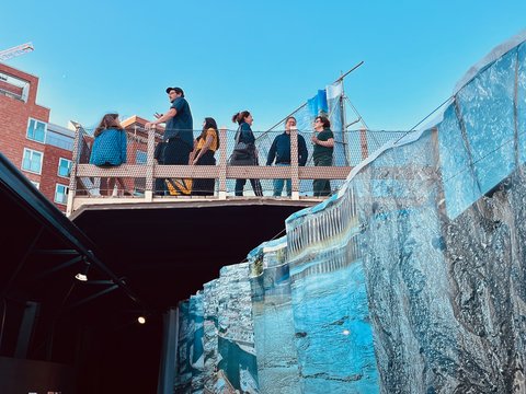 "Etwa 10 Personen in sommerlicher Kleidung stehen auf einer Plattform neben einer blauen Hafenmauer. Sie betrachten gemeinsam großformatige architektonische Pläne und Skizzen. Im Hintergrund sind ein moderner Backsteinbau und das Hafenbecken sichtbar. Die Sonne scheint bei klarem blauen Himmel."