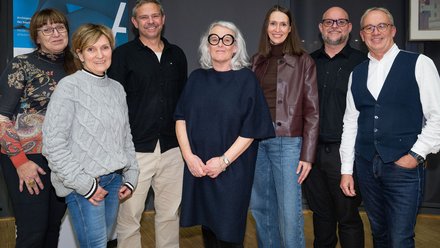 Eine Gruppe von acht erwachsenen Personen steht eng nebeneinander in einem Innenraum vor einer dunklen Wand und einem aufgestellten Banner, sie tragen überwiegend legere bis halbformelle Kleidung und blicken ruhig in die Kamera, wodurch das Bild ein offizielles Gruppenfoto eines Vorstands oder Gremiums zeigt und für gemeinschaftliche Verantwortung, institutionelle Zusammenarbeit und die öffentliche Vertretung von Baukultur steht.
